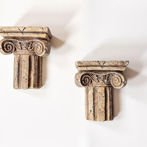 Pode incluir: Dois capitéis de coluna decorativos com aparência envelhecida. Os capitéis são de cor bege-castanho com volutas intrincadas e ranhuras verticais. Estão sobre um fundo branco.