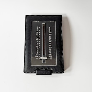 Puede incluir: Un directorio negro vintage con un dial deslizante plateado y una cubierta de cuero negro. El dial tiene letras de la A a la Z. El directorio es rectangular y tiene una peque&ntilde;a solapa en la parte inferior.