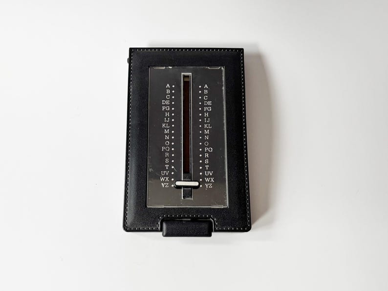 Puede incluir: Un directorio negro vintage con un dial deslizante plateado y una cubierta de cuero negro. El dial tiene letras de la A a la Z. El directorio es rectangular y tiene una peque&ntilde;a solapa en la parte inferior.