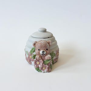 Puede incluir: Una caja de baratijas de cerámica con tapa, con un diseño de panal de abeja gris claro. Un oso de peluche marrón está adherido a la parte delantera, rodeado de flores rosas y hojas verdes. La caja se utiliza probablemente para guardar objetos pequeños.