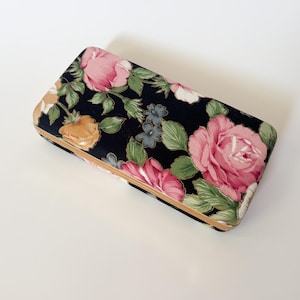 Pode incluir: Um estojo rígido retangular com exterior em tecido preto e um padrão floral de rosas cor-de-rosa, folhas verdes e pequenas flores amarelas e azuis. O estojo possui uma moldura de metal dourado nas bordas e um mecanismo de fecho articulado.
