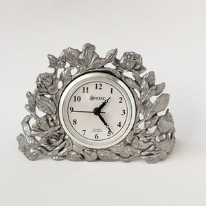 May include: A silver-coloured quartz clock featuring a rose floral design. The clock face is white with black numerals and hands. The brand name "Seagull" is printed on the clock face. A decorative item.