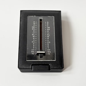 Puede incluir: Un directorio negro vintage con un selector de letras deslizante. El libro tiene forma rectangular con una cubierta texturizada similar al cuero y una ventana de pl&aacute;stico transparente que muestra el alfabeto. El selector est&aacute; en el centro.