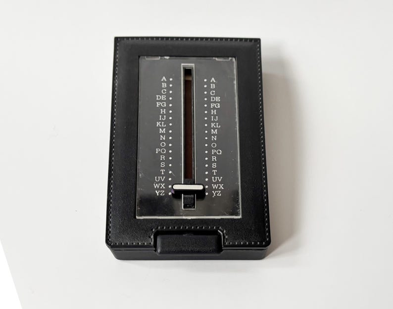 Puede incluir: Un directorio negro vintage con un selector de letras deslizante. El libro tiene forma rectangular con una cubierta texturizada similar al cuero y una ventana de pl&aacute;stico transparente que muestra el alfabeto. El selector est&aacute; en el centro.