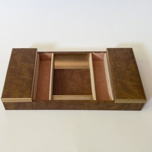 May include: A rectangular brown jewelry box with a textured finish and gold trim. The interior features multiple compartments lined with a pinkish-beige fabric. The box is open, revealing the storage space.
