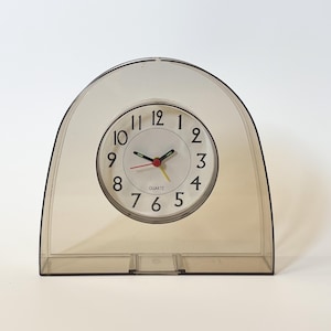 Puede incluir: Un reloj analógico de cuarzo con un marco transparente y arqueado. La esfera del reloj es blanca con números y manecillas negras. El segundero es amarillo, y las manecillas de minutos y horas son negras. El reloj mide aproximadamente 15 cm de ancho y 13 cm de alto.