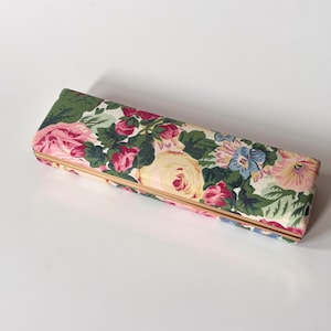 Peut inclure: Étui à lunettes rectangulaire recouvert d'un tissu floral. Le motif présente des roses roses, des pensées bleues et des feuilles vertes sur un fond crème. Les bords sont bordés de métal doré.