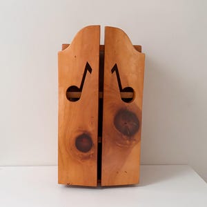 以下が含まれることがあります： 2つのドアが付いた木製の壁掛けキャビネット。キャビネットは明るい茶色の木材で作られており、各ドアに音符の切り抜きがあります。ドアは閉じており、キャビネットは素朴で手作りの外観をしています。
