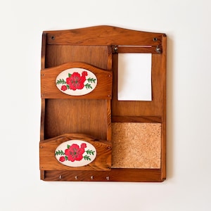 May include: Wooden wall-mounted organizer with two mail slots featuring oval floral designs with red flowers. Includes a corkboard, a space for a notepad, and three hooks for keys. The organizer is made of wood.