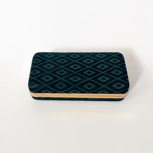 May include: A rectangular clutch bag featuring a dark green velvet exterior with a repeating black diamond pattern. The bag is finished with a thin, metallic gold-toned frame along the center edge, providing a structured and clean appearance.