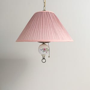 Peut inclure: Une lampe suspendue dotée d'un large abat-jour en tissu plissé rose clair. Sous l'abat-jour, une pièce décorative en verre blanc sphérique est ornée d'un motif floral peint en rose, vert et bleu. Une chaînette métallique est suspendue.