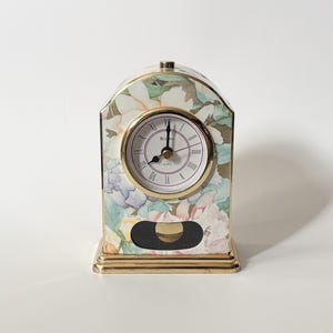 May include: A Bulova quartz mantel clock with an arched top and a gold-tone metal base. The clock face features Roman numerals and black hands, set against a floral pattern in soft pastel shades of pink, blue, and green on the exterior casing.
