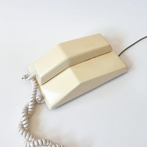 Puede incluir: Teléfono vintage color crema con un cable de auricular en espiral y un cable de alimentación recto. El teléfono tiene un diseño geométrico con una base rectangular y una sección superior elevada. El teléfono está sobre un fondo blanco.