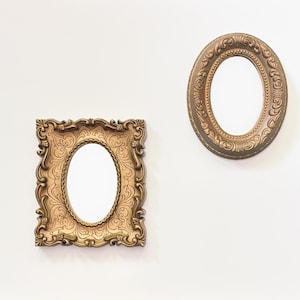 May include: Two wall mirrors with ornate, metallic gold-toned frames. One mirror is rectangular with rounded corners and an oval center, while the other is a simple oval shape. Both frames feature intricate, raised scrollwork and decorative patterns.