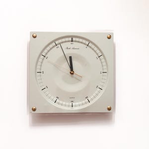 Puede incluir: Un reloj de pared cuadrado, gris claro, con una esfera circular. El reloj tiene manecillas y marcadores negros, con detalles dorados en las esquinas. El texto "Park Avenue" es visible en la esfera.