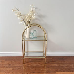 Puede incluir: Una estantería arqueada de metal dorado con dos estantes de cristal transparente. El estante superior sostiene un jarrón de cerámica blanca acanalada con follaje plateado seco, junto a dos botellas pequeñas de cristal.
