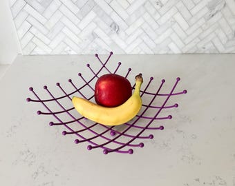 Può includere: Un cestino di frutta in filo di ferro viola con una mela rossa e una banana gialla. Il cestino ha un design geometrico con linee intersecanti e piccoli accenti a forma di sfera. La frutta è fresca e matura.