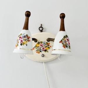May include: A white wall sconce with two bell-shaped shades featuring floral designs. The shades are painted with a bouquet of pink, yellow, and red flowers. The sconce has a white base and a brown wooden stem.