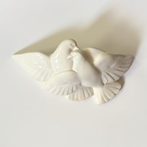 Peut inclure: Une plaque murale en céramique blanc cassé brillant représentant deux colombes sculptées en plein vol. Les oiseaux sont placés côte à côte, ailes déployées, avec des plumes détaillées et des formes lisses et arrondies.