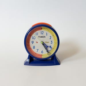 May include: A blue Timex analogue alarm clock with a round face featuring a white centre, a red left half, and a yellow right half. The clock has blue numbers, a green hour hand, a black minute hand labelled 'Minute', and an orange top button.