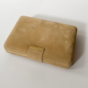 May include: A rectangular tan suede clutch bag featuring a smooth, soft-touch exterior surface. The bag is secured with a central vertical strap that includes a polished gold-tone metal accent piece at the closure point, providing a clean finish.