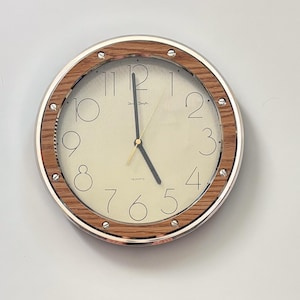 May include: A round wall clock featuring a light cream-coloured face with thin black numerals from one to twelve. The clock has a wood-grain patterned frame accented by a silver metallic outer rim and six visible silver screws. Black hands indicate the time.