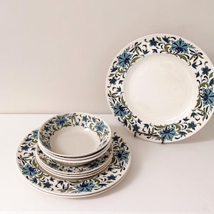 May include: A set of vintage white ceramic dinnerware with a blue and green floral design. The set includes dinner plates and smaller bowls, perfect for serving meals. The plates are arranged on a white surface.