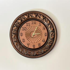 May include: A round wall clock with a metallic copper finish. The clock face features a central sunburst pattern, raised numerals from 1 to 12, and thin gold-coloured hands. The outer border is decorated with a relief pattern of acorns and leaves.