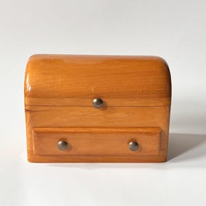 May include: A small wooden chest with a curved top and a drawer. The chest is made of light brown wood and has two small, round, dark metal knobs. The chest is likely used for storing jewellery or small items.