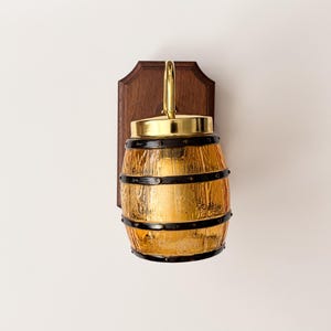 May include: A wall-mounted light fixture with a barrel-shaped amber glass shade. The fixture has black metal bands, a gold-colored top, and a brass handle. It is mounted on a dark wood backing.