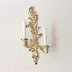 May include: A wall-mounted candle sconce featuring an off-white finish with gold-toned accents. The fixture has an ornate, scrolling leaf design and two candle holders, each containing a plain white taper candle. The structure is vertical.