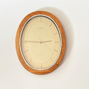 May include: An oval wall clock featuring a light brown wood frame and a silver-toned metal bezel. The clock face is cream-colored with thin gold-toned hour markers, gold-toned hands, and the word Sunbeam printed in red near the top center.