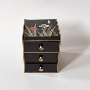 Peut inclure: Une boîte à bijoux noire à trois tiroirs avec des bordures et des boutons dorés. Le dessus de la boîte présente un motif floral avec des accents orange, blancs et verts. La boîte est rectangulaire et repose sur une surface blanche.