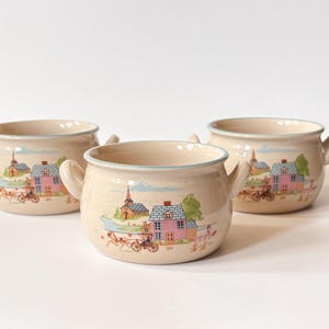 Peut inclure: Trois bols en céramique de couleur crème, chacun doté de deux anses latérales. L'extérieur présente une illustration colorée d'une maison rose, d'une église et d'une calèche. Les bords sont soulignés d'un fin liseré bleu et la finition est lisse.