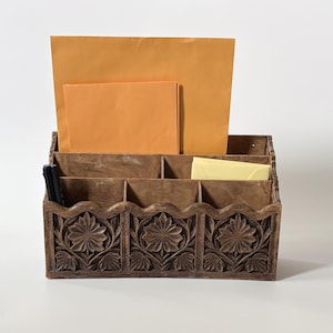 May include: A brown wooden desk organiser featuring three carved floral panels on the front and a scalloped top edge. The organiser has multiple compartments holding large orange envelopes, a smaller yellow envelope, and two black pens.