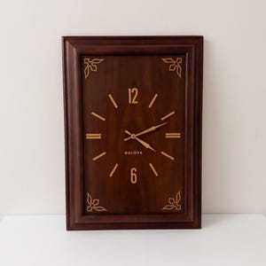 May include: A rectangular Bulova wall clock with a dark brown wooden frame and face. The clock features gold-coloured hands and numerals, with the number 12 at the top and 6 at the bottom. Decorative floral accents are in each corner.
