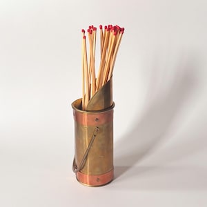 May include: A brass and copper match holder filled with wooden matches. The cylindrical holder has a handle and is accented with copper bands and rivets. The matches have red tips and are arranged upright.