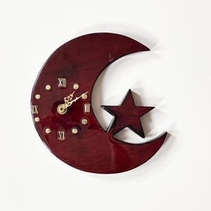 May include: A burgundy-colored wall clock in the shape of a crescent moon with a star accent. The clock face features Roman numerals and gold-colored hands. Decorative gold-colored dots are placed around the clock face.