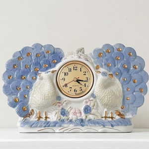 Puede incluir: Un reloj de cerámica con diseño de pavo real. Cuenta con una esfera dorada, dos aves blancas y detalles de plumas azules con detalles dorados. El reloj mide aproximadamente 20 cm de ancho y 15 cm de alto.