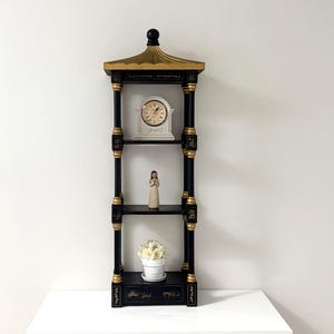 Peut inclure: Une grande étagère de présentation noire et dorée de style pagode à trois niveaux. L'étagère supérieure contient une horloge analogique blanche, le niveau central présente une petite figurine debout et le niveau inférieur contient une plante en pot blanche.
