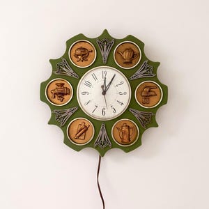 May include: A round olive green wall clock with a central white face featuring black numerals and hands. The frame is decorated with six circular copper-toned relief plaques depicting kitchen items, including a teapot, grinder, and rolling pin.