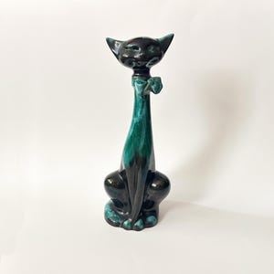 May include: A tall, stylized ceramic cat figurine featuring a glossy finish with a blend of deep black and vibrant teal colors. The cat has an elongated neck, pointed ears, and a small bow detail around its neck, sitting in an upright posture.