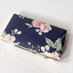 Peut inclure: Une pochette rectangulaire avec un fond bleu marine et un motif floral avec des fleurs roses et blanches et des feuilles vertes. La pochette a une bordure dorée.
