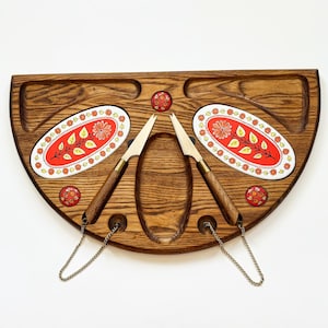 May include: A semi-circular wooden serving board featuring two oval ceramic inlays with red floral patterns, three small circular decorative accents, and two metal cheese knives with wooden handles attached by thin silver-toned chains.