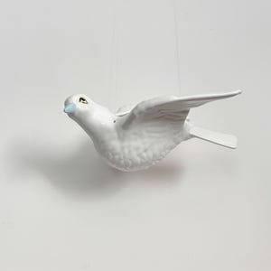 May include: A white ceramic bird ornament with a glossy finish, featuring a light blue beak and detailed wing feathers. The bird is depicted in a flying position with wings extended, suspended by thin, nearly invisible strings against a plain background.