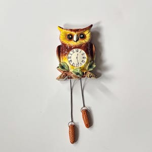 May include: A ceramic owl-shaped clock with a brown and yellow color scheme. The clock face has Roman numerals and is surrounded by a decorative branch with green leaves. Two decorative pinecone-shaped weights hang below.