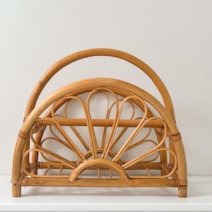 Może przedstawiać: Naturalny, rattanowy stojak na czasopisma. Stojak posiada zakrzywiony uchwyt i dekoracyjny wzór promieni słonecznych z przodu. Element wykonany jest z plecionego rattanu i nadaje się do przechowywania czasopism lub książek.