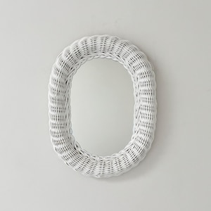 May include: White oval wicker mirror. The mirror has a woven frame and a reflective surface. The frame is made of tightly woven wicker and is painted white. The mirror is a decorative wall hanging.