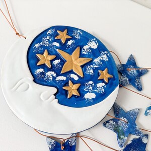 Op de afbeelding: Een blauwe en witte keramische maan met gouden sterren die hangt aan een koperen draad met blauwe stervormige ornamenten.