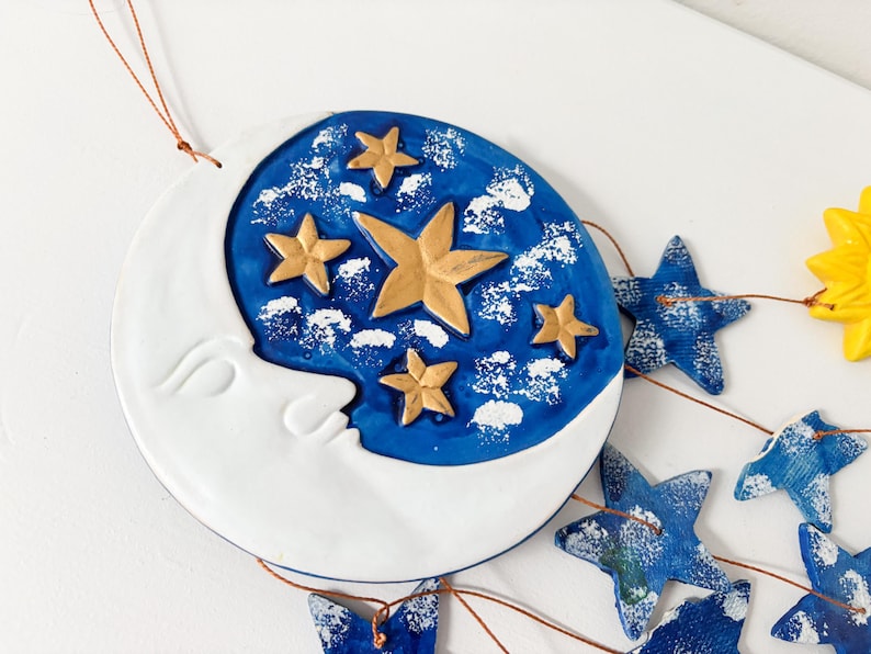 Op de afbeelding: Een blauwe en witte keramische maan met gouden sterren die hangt aan een koperen draad met blauwe stervormige ornamenten.